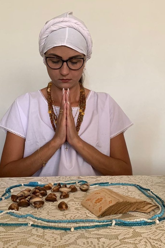 Gabriela em prece, de mãos unidas, diante de búzios, cartas e guias azuis e brancas sobre a mesa