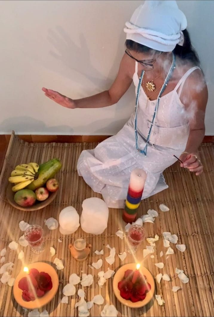 Gabriela de branco, sentada sobre uma esteira, preparando um ritual com velas, rosas vermelhas, pétalas brancas e frutas