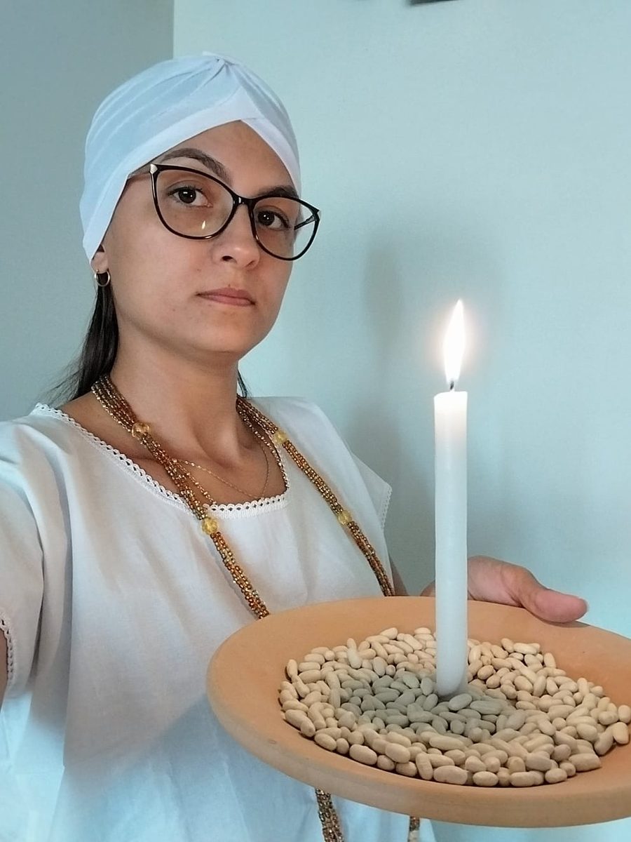 Astróloga Gabriela de turbante branco e óculos, segurando uma vela branca acesa sobre um alguidar com feijão